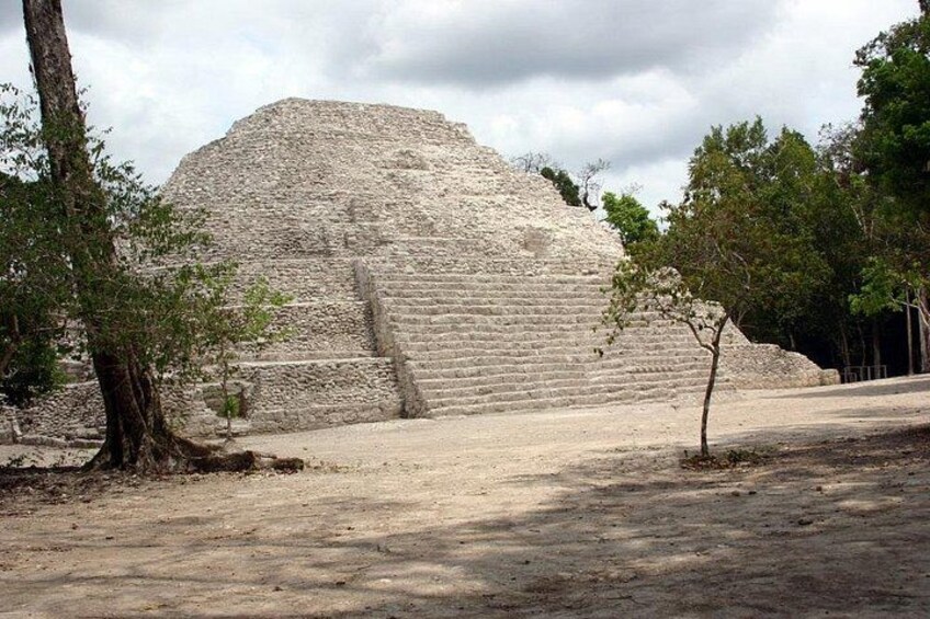 A large stone pyramid with a staircase leading up to its entrance, surrounded by trees and a clear sky. 1