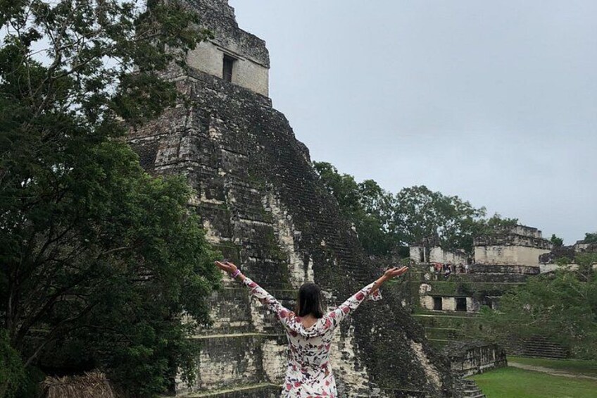 Una persona con los brazos levantados frente a una gran estructura piramidal antigua rodeada de árboles. 2