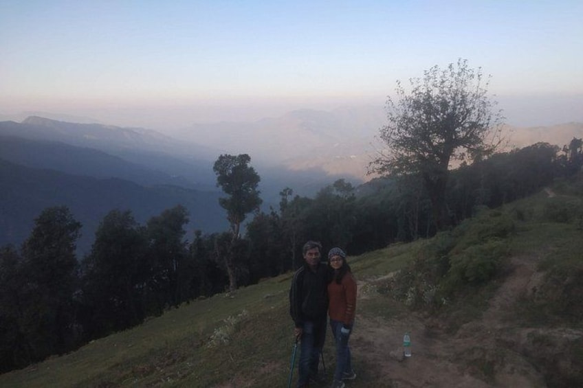 Two people standing on a grassy hill with a view of distant mountains and a tree. 5