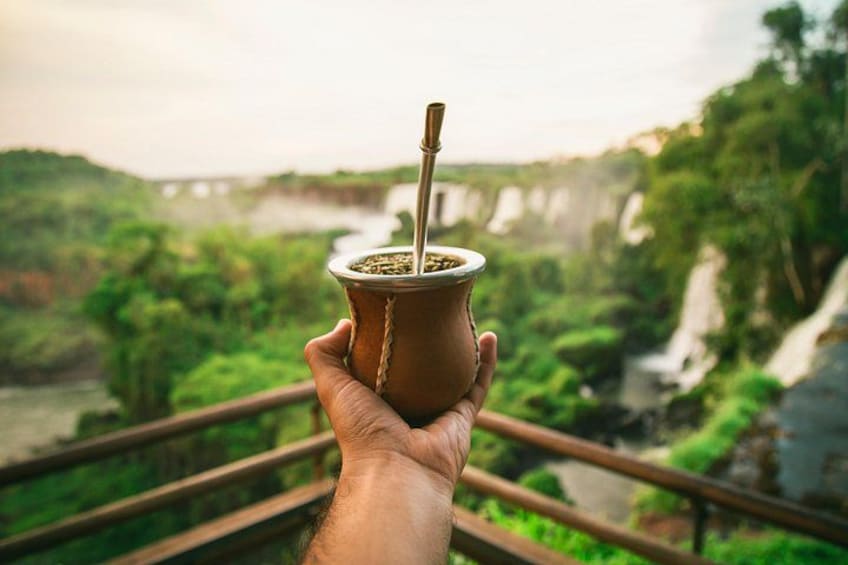 Una mano sorregge un recipiente tradizionale sudamericano con una cannuccia, con vista su un paesaggio verdeggiante con un fiume e una cascata. 2