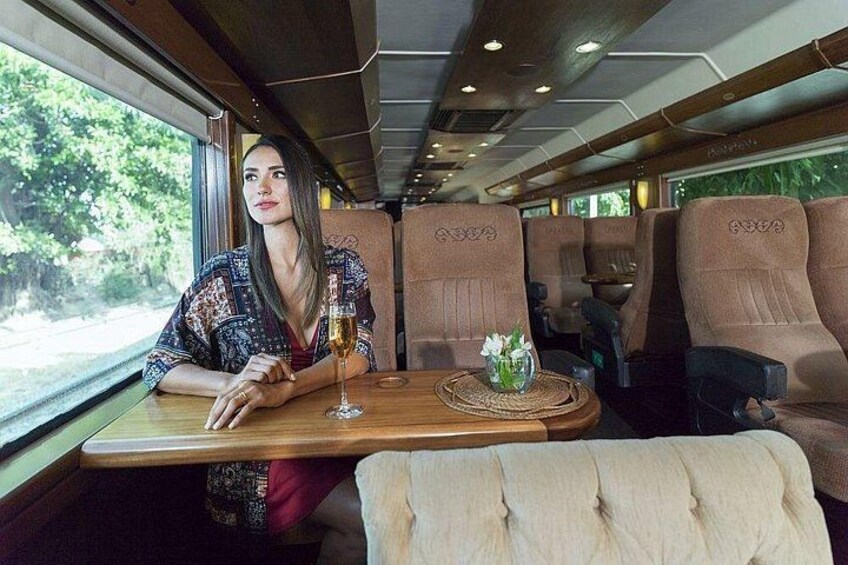 Ein Zuginnenraum, in dem eine Frau an einem Tisch Platz nimmt, mit einem Weinglas und einer verzierten Platte. 3