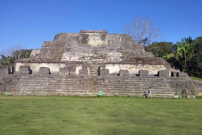 A large ancient pyramid with a stepped structure and a flat top, surrounded by a grassy area with a few people walking around. 1