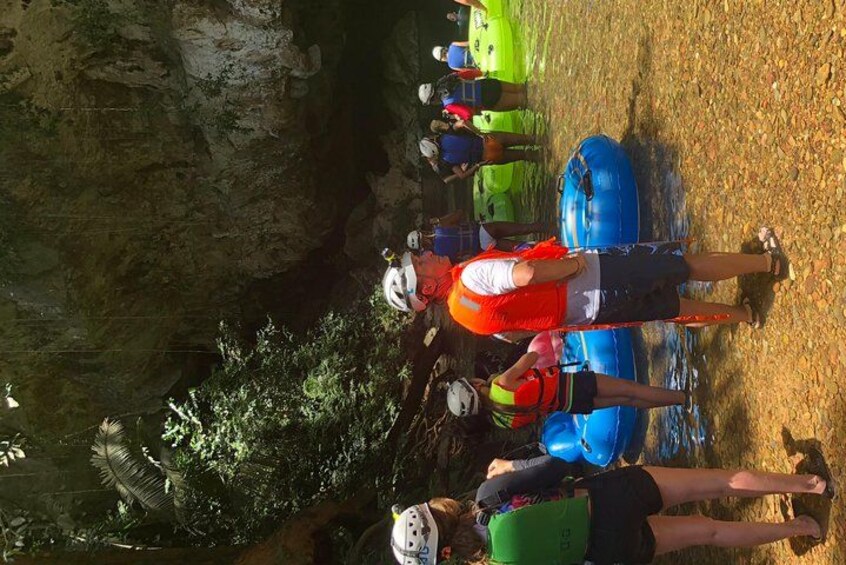 A group of people standing near a cave entrance, some wearing helmets and life vests, with inflatable rafts in the background. 2