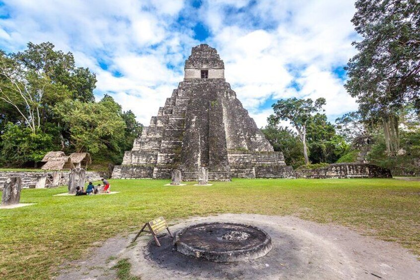 A large ancient pyramid surrounded by lush greenery and a small structure with people nearby. 3