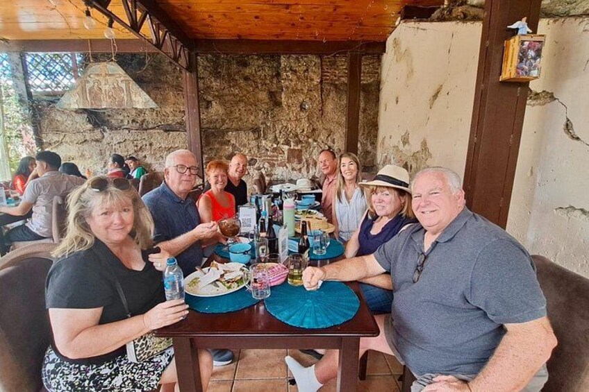A group of people seated around a table with food and drinks, in an outdoor setting with stone walls and wooden beams. 3