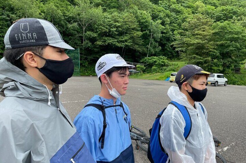 Three people standing outdoors, wearing jackets and backpacks, with trees and a parked vehicle in the background. 5