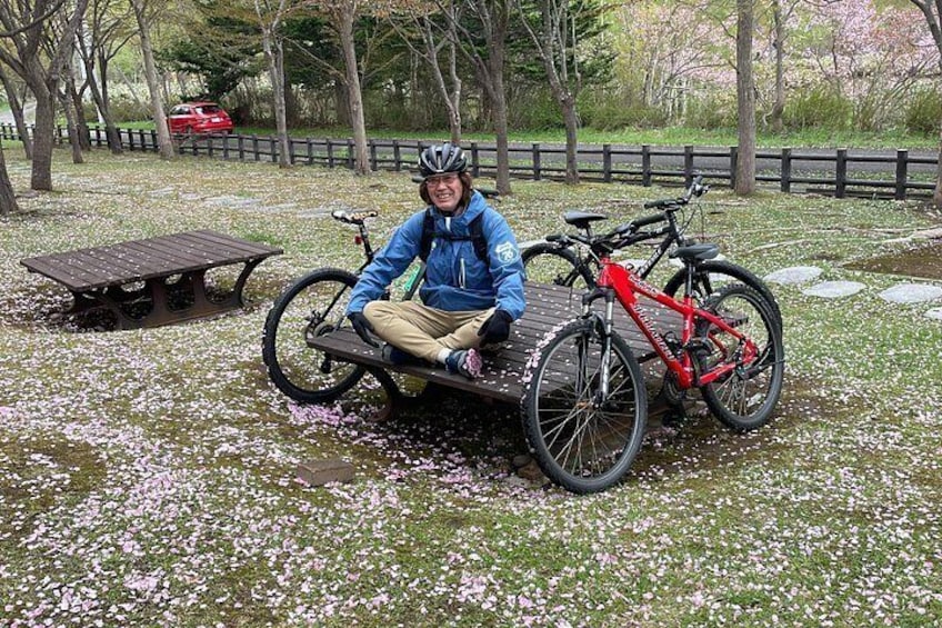 A person sitting on a bench with a bicycle, surrounded by cherry blossoms. 2