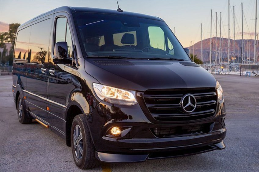 Un furgone Mercedes di colore scuro parcheggiato in un parcheggio, con uno sfondo che mostra un porto turistico e imbarcazioni. 3