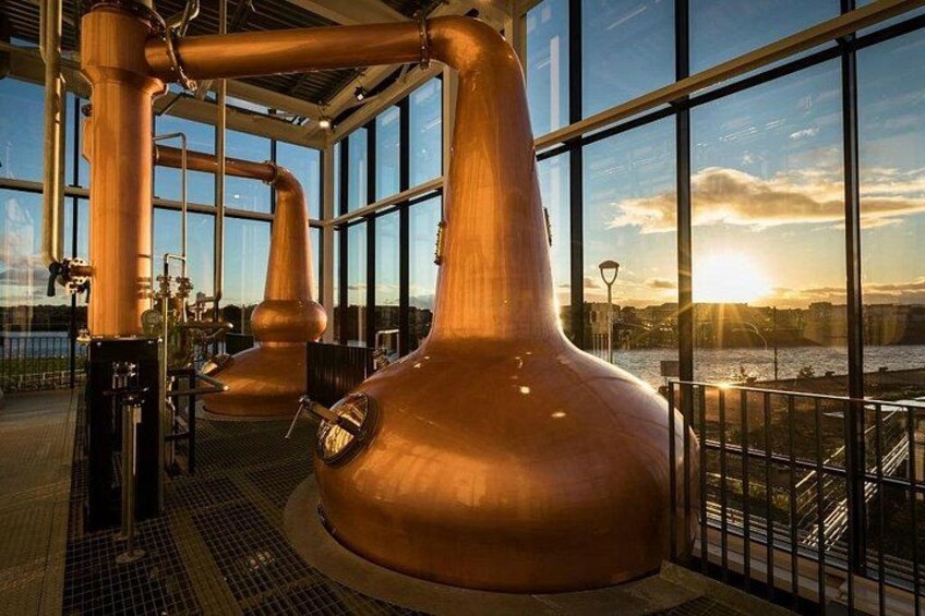 A distillery with copper stills and a view of a river at sunset. 4