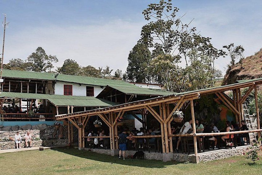 Un área de estar al aire libre con asientos de madera y techo de paja, rodeada por un campo cubierto de hierba y un edificio con techo verde. 4