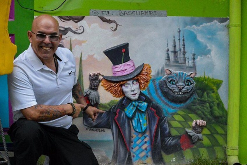 A man sitting next to a colorful mural with a cat and a figure in a top hat. 2