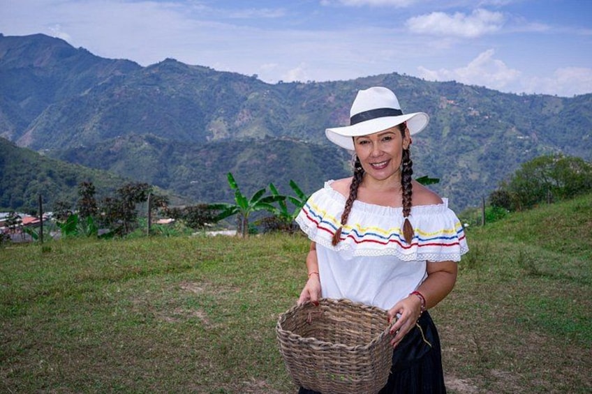 A person holding a woven basket, wearing a white hat and a colorful off-shoulder blouse, standing on a grassy hill with mountains in the background. 5