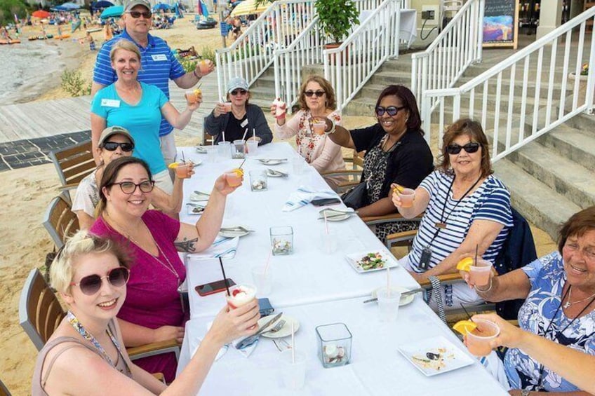A group of people seated at a table outdoors, with a beach and staircase in the background. 4