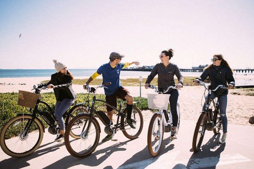 Fyra personer cyklar på en strand med en brygga i bakgrunden. 1