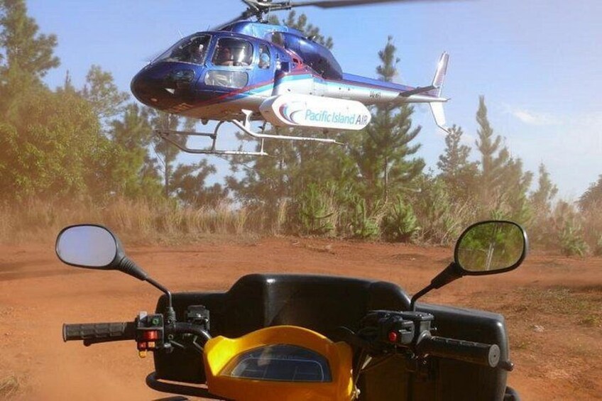 A helicopter with 'Pacific Island Air' written on it flying above a dirt road with a yellow vehicle parked on the side. 4