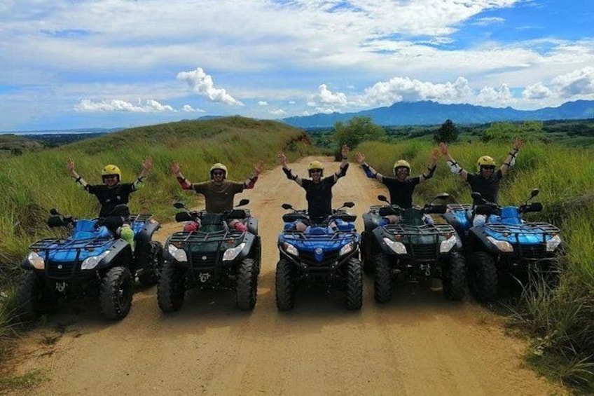 Four people on ATVs on a dirt road with a grassy landscape and mountains in the background. 3