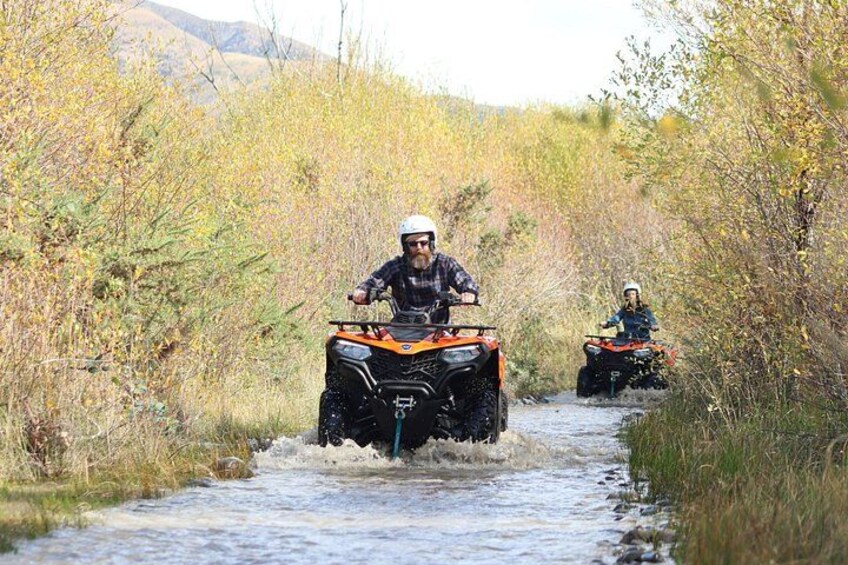 Two people riding ATVs on a narrow water crossing surrounded by dense vegetation. 2