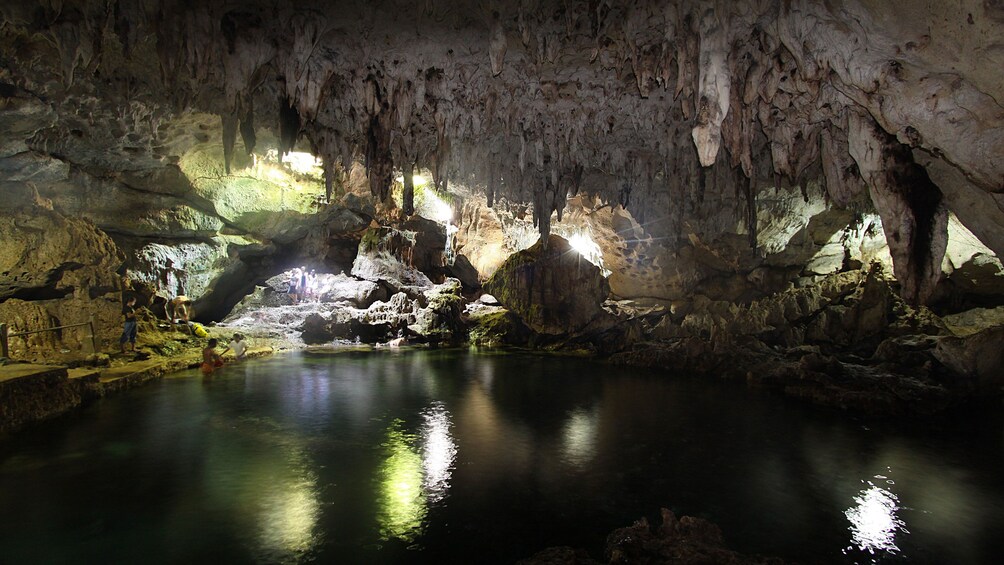 A cave with stalactites, a water pool, and people exploring. 5
