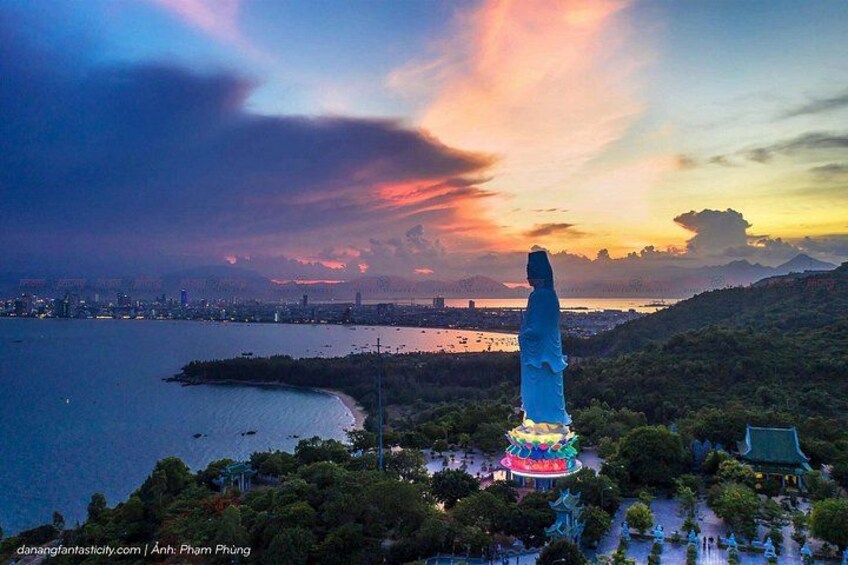 A tall, illuminated monument against a sunset sky, surrounded by greenery and a coastal cityscape. 2