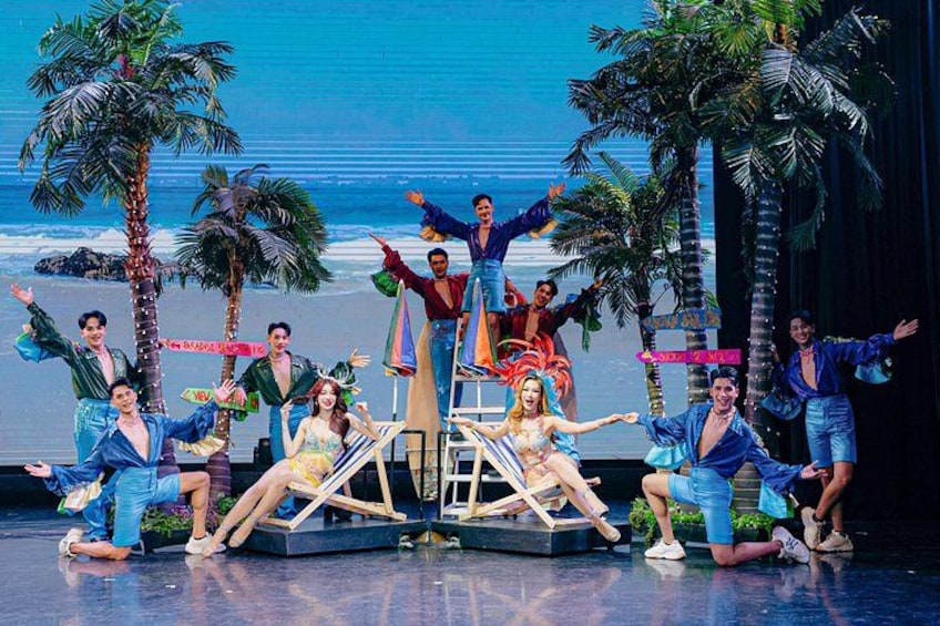 Une scène où des artistes, vêtus de costumes, se produisent, avec des palmiers et un décor représentant une plage en arrière-plan. 2