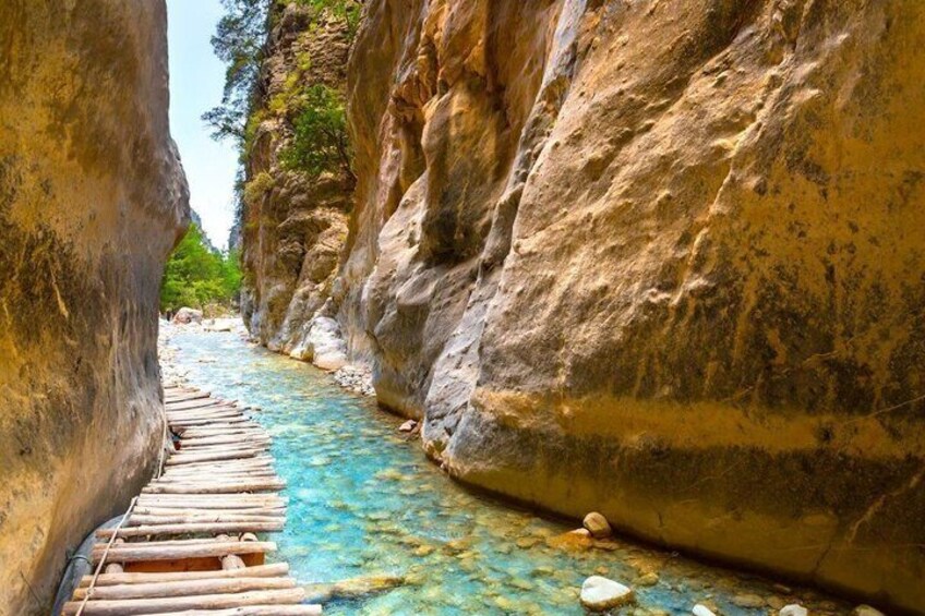 A narrow canyon with a clear blue river, wooden planks along the riverbed, and rocky cliffs. 4