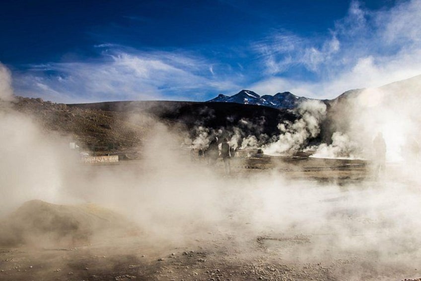 A geothermal area with steam rising from the ground, a clear sky, and a mountain range in the background. 3