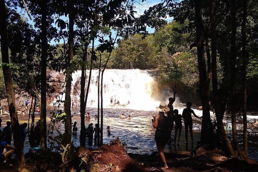 A waterfall in a forested area with people standing and sitting around the water. 1