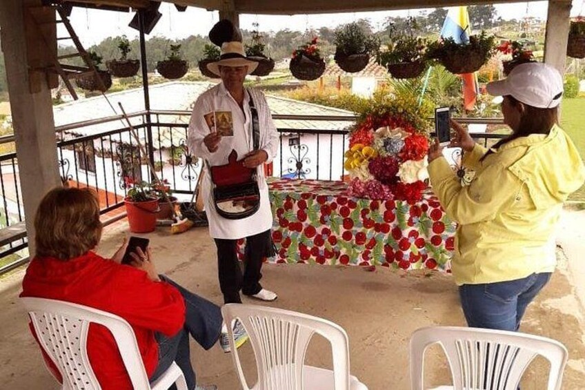 A person in a white hat and apron standing on a porch with a floral arrangement, a person taking a photo, and another person seated. 5