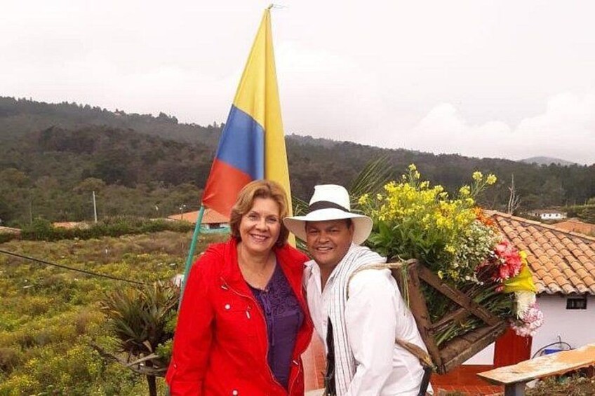 Two people standing in a rural area with a flag, flowers, and a house with a red roof. 4