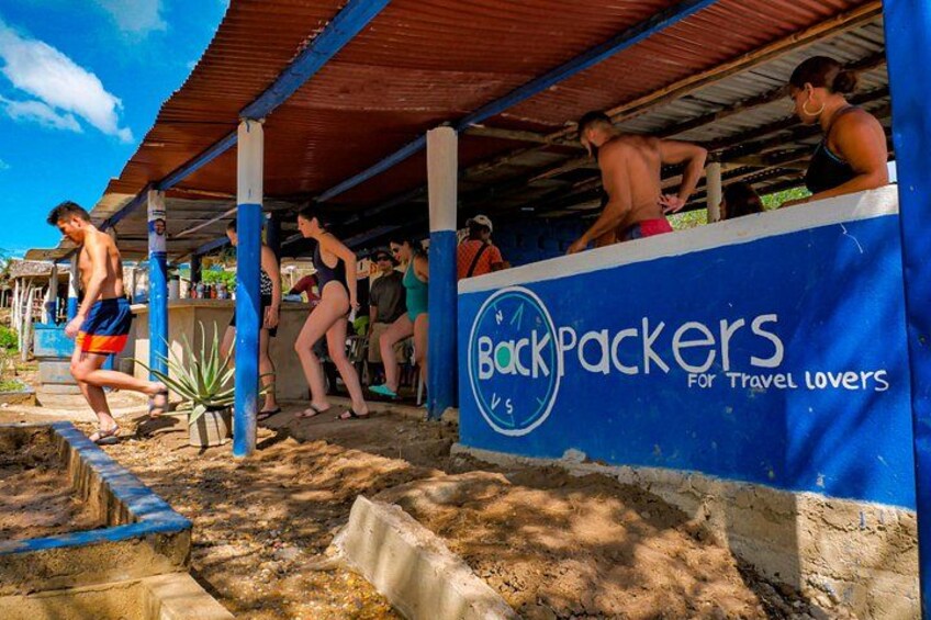 A beachside area with a covered structure, people engaging in water activities, and a sign reading 'Backpackers for Travel Lovers'. 2