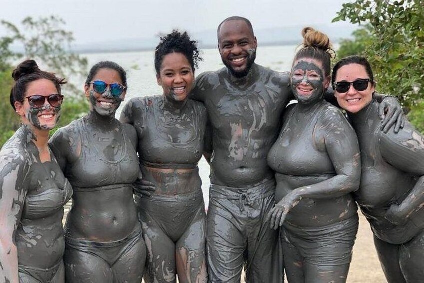 A group of people covered in mud, standing on a beach with a body of water in the background. 1