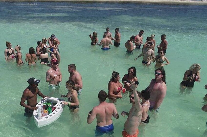 Un groupe de personnes se trouve dans une étendue d’eau peu profonde et claire. Certaines sont debout, d’autres sont assises, et un petit bateau est visible au premier plan. 2
