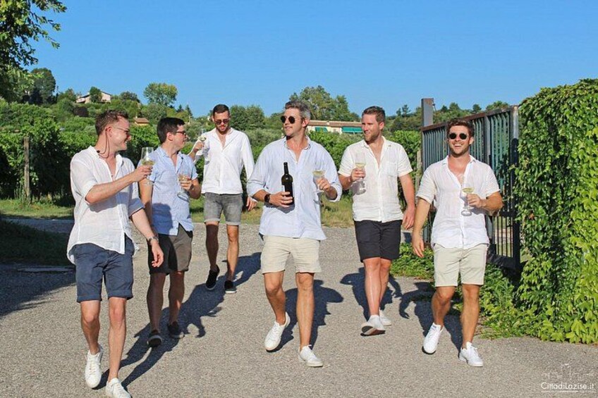 Een groep mensen loopt over een pad, ze houden drankjes vast, met groen en een hek op de achtergrond. 4