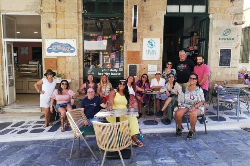 A group of people sitting and standing outside a café with a sign that says "EXPLORE ANDROS." 1