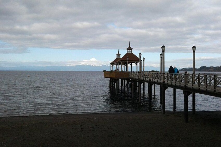 Une jetée s’avançant dans une eau calme, agrémentée d’un belvédère et de lampadaires, une montagne enneigée visible au loin et un ciel nuageux. 3