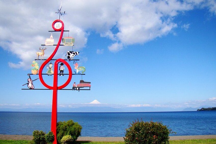 Un symbole de note de musique rouge, agrémenté de divers emblèmes culturels, sur un fond de ciel dégagé et de mer calme. 1