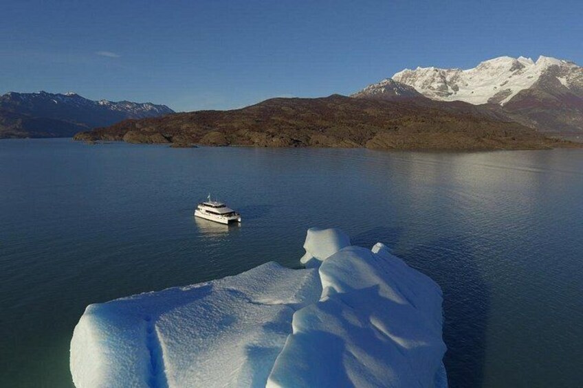 A boat on calm water, a large iceberg, and snow-capped mountains in the background. 1
