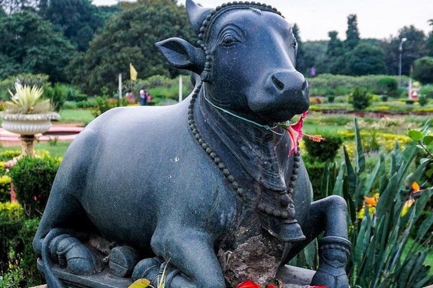 A statue of a cow in a garden with greenery and a fountain in the background. 4
