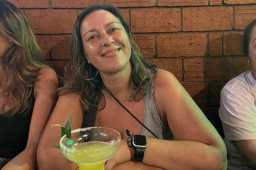 Una persona sorseggia un cocktail guarnito con una fetta di lime, indossa uno smartwatch e siede a un tavolo con una parete di mattoni sullo sfondo. 4