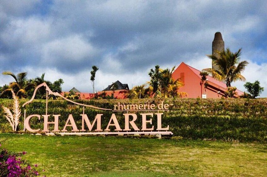 A sign with the text 'rhumerie de CHAMAREL' in a lush garden setting with a red building and palm trees in the background. 4