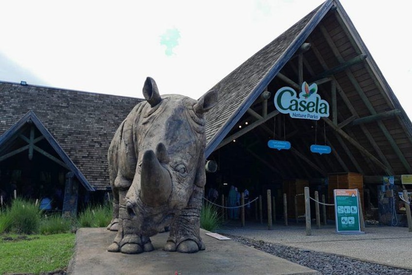A large rhino statue in front of a building with a pointed roof and a sign that reads 'Casela Nature Park'. 1