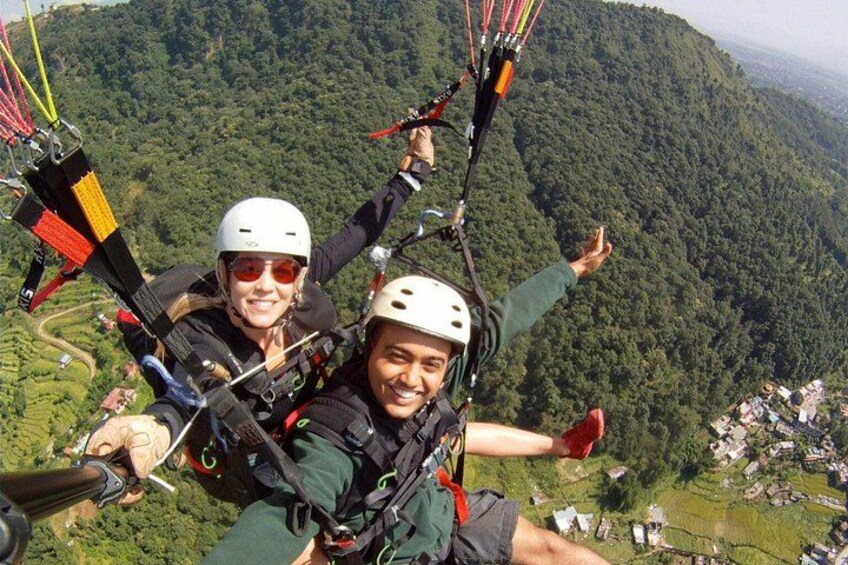 Two people paragliding over a mountainous landscape with lush greenery and a small village below. 2
