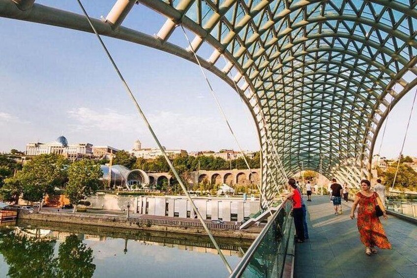 Een voetgangersbrug met een gebogen metalen dak, een rivier eronder en gebouwen op de achtergrond. 3