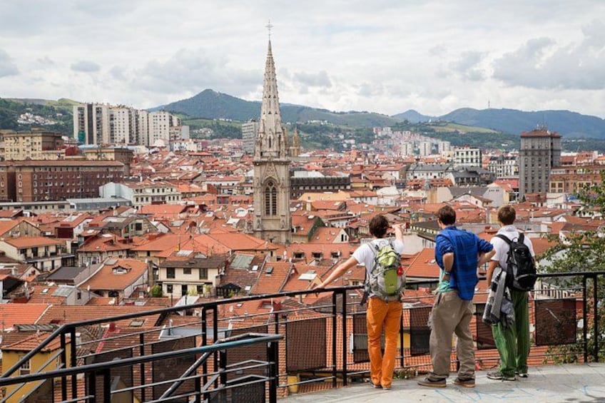 Eine Gruppe von Personen steht auf einem Balkon mit Blick auf eine Stadtlandschaft, geprägt von einem markanten Kirchturm. 3