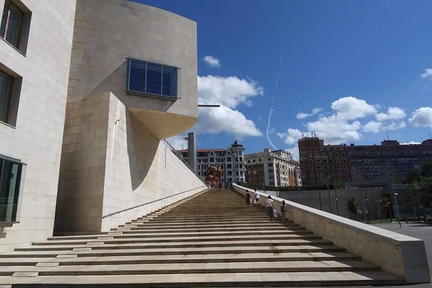 A staircase leading up to a modern building with a unique architectural design, a clear sky, and a distant view of traditional buildings. 4