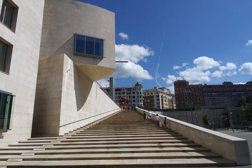 Un bâtiment moderne doté d’une architecture originale, un escalier menant à l’entrée et un ciel bleu azur parsemé de nuages. 2