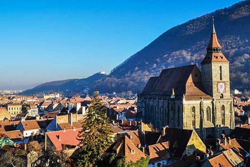 Una cittadina con una chiesa imponente, caratterizzata da un'alta torre e un orologio, circondata da edifici con tetti rossi e una montagna sullo sfondo. 3