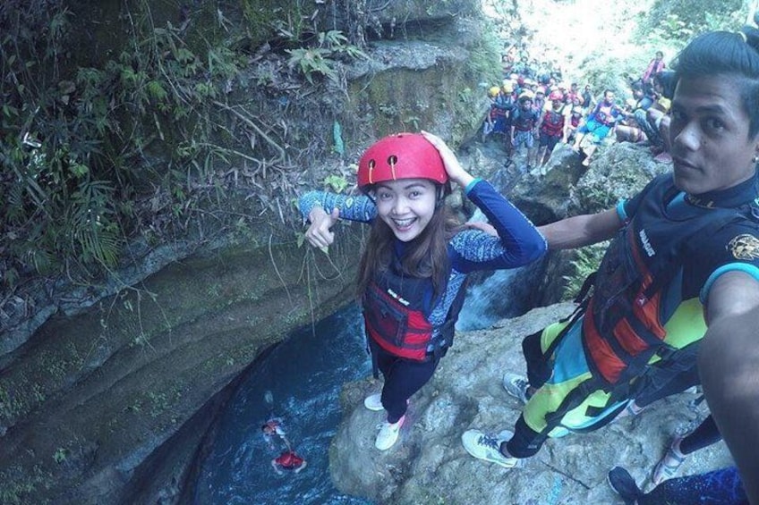 A person in a red helmet and blue jacket standing on a rock near a water body, with a group of people in the background. 4