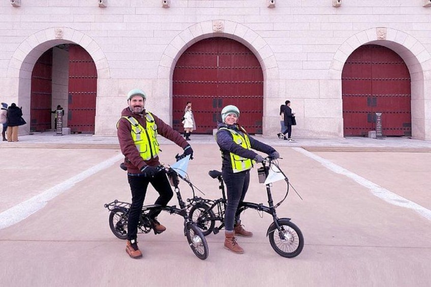 Two people on bicycles in front of a building with large red doors. 2