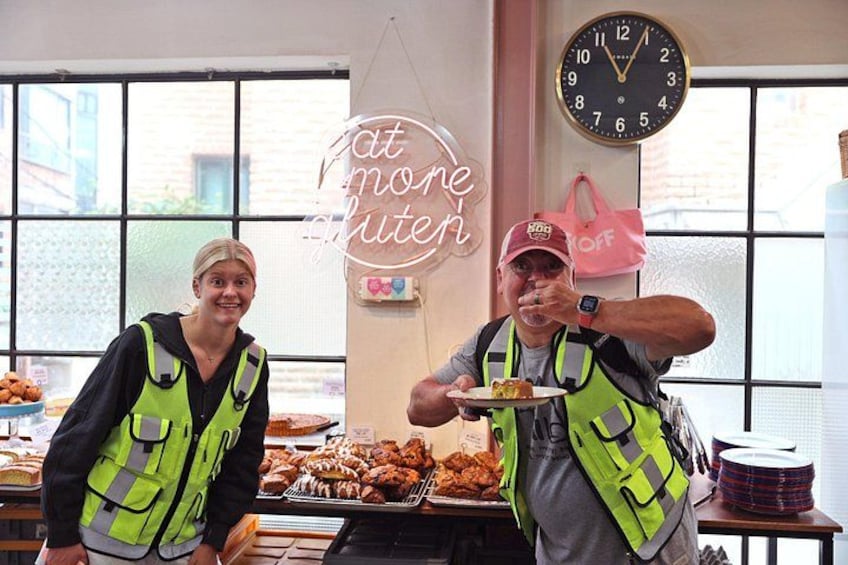 Two people in a bakery wearing safety vests, a clock, and a neon sign. 3